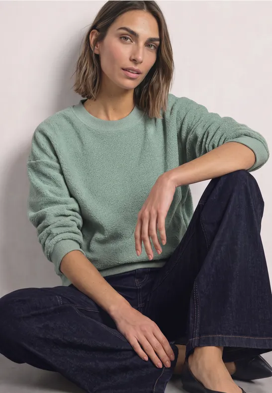 Eine Frau sitzt im Schneidersitz, trägt grünen Pullover und weite, dunkle Jeans.