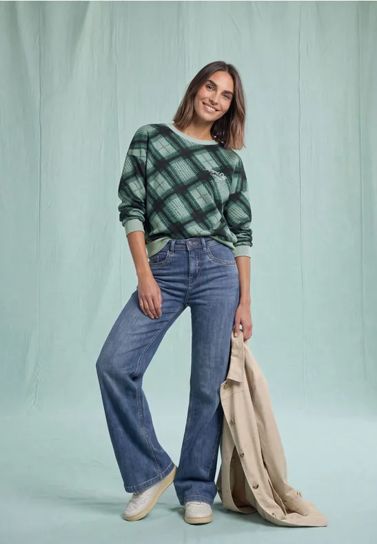 Femme en haut à carreaux verts, jean flare bleu, baskets blanches, manteau beige à la main devant un fond vert.
