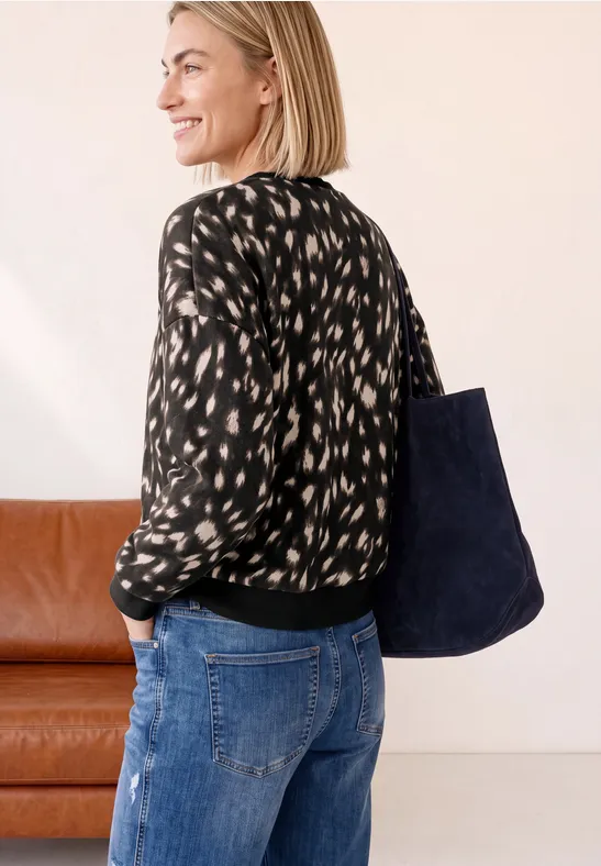 Frau in gemusterter Bluse und Jeans trägt eine große blaue Tasche, neben einem braunen Sofa.