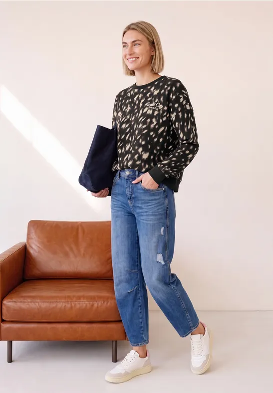 Frau mit schwarzem gemustertem Pullover, blauen Jeans und weißen Sneakern steht neben einem braunen Ledersofa.