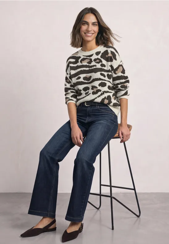Femme assise sur un tabouret, en pull imprimé animal, jean bleu foncé et escarpins noirs, souriante.