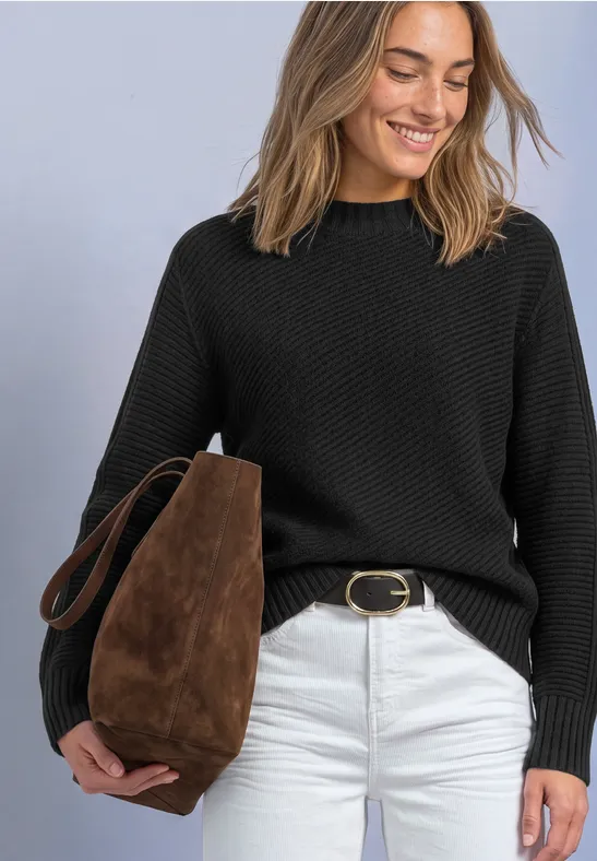 Frau mit schwarzem Strickpullover, weißer Hose und brauner Suede-Tasche, lächelt und blickt nach unten.