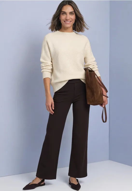 Frau in cremefarbenem Pullover, dunkelbrauner Hose und brauner Tasche vor schlichtem Hintergrund.