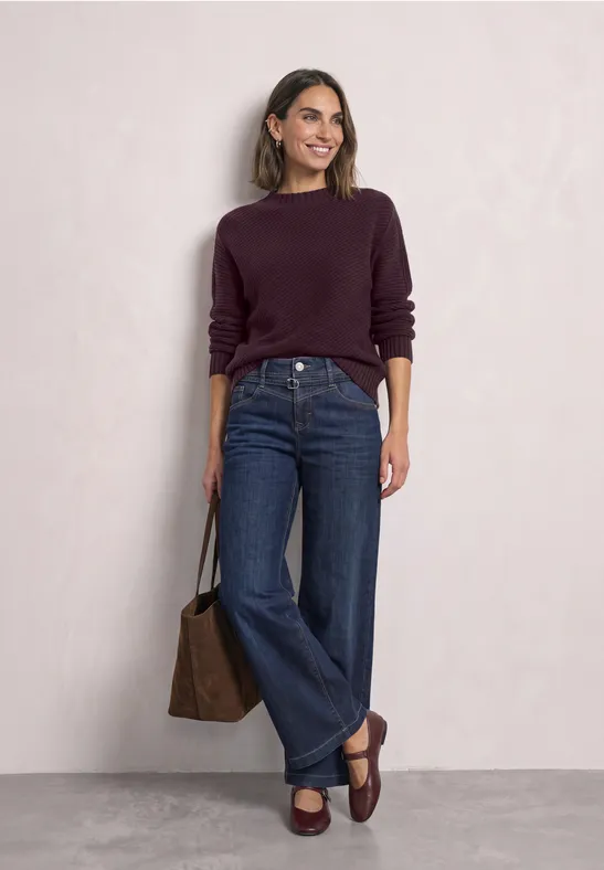 Frau in lila Pullover, weitem Jeans und braunen Loafers mit passender Tasche.