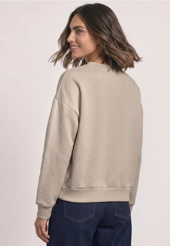 Frau im beigen Sweatshirt mit langen Ärmeln und blauen Jeans, Rückansicht.