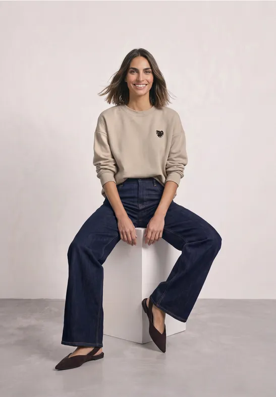 Frau mit beiger Sweatshirt und weiten Jeans sitzt lächelnd auf einem weißen Hocker vor hellem Hintergrund.