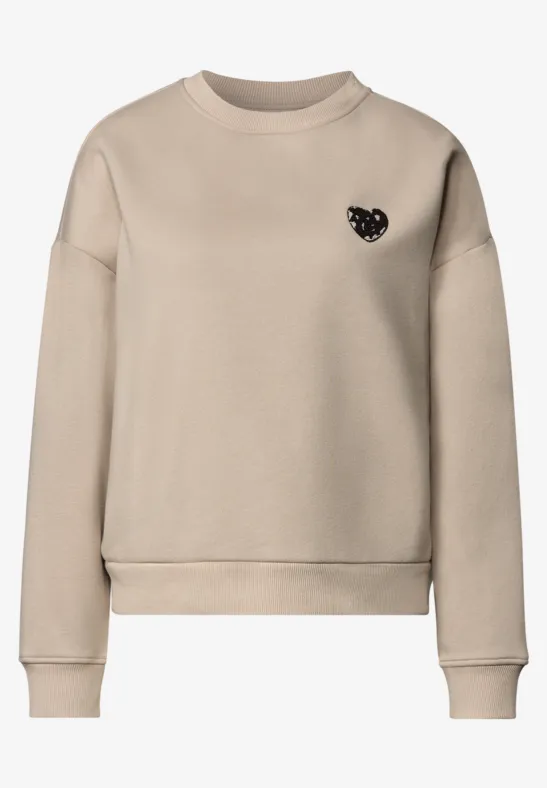 Beiges, schlichtes Sweatshirt mit Rundhalsausschnitt und kleinem schwarzen Herz-Emblem auf der Brust.