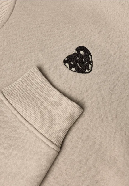 Beiges Sweatshirt mit aufgesetzter Tasche und kleinem schwarzen Herzpatch mit weißen Dreiecksmustern.