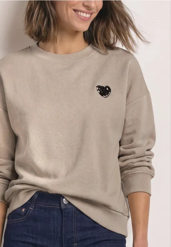 Eine Frau trägt einen beigen Pullover mit kleinem schwarzem Katzen-Motiv und eine Jeans.