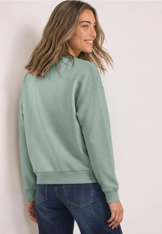 Eine junge Frau in hellgrünem Pullover schaut lächelnd über die Schulter.