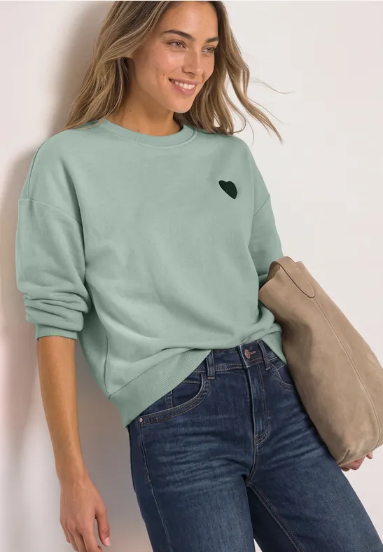 Frau in grünem Pullover mit Herz-Logo und Jeans, hält beige Jacke, lächelt vor hellem Hintergrund.