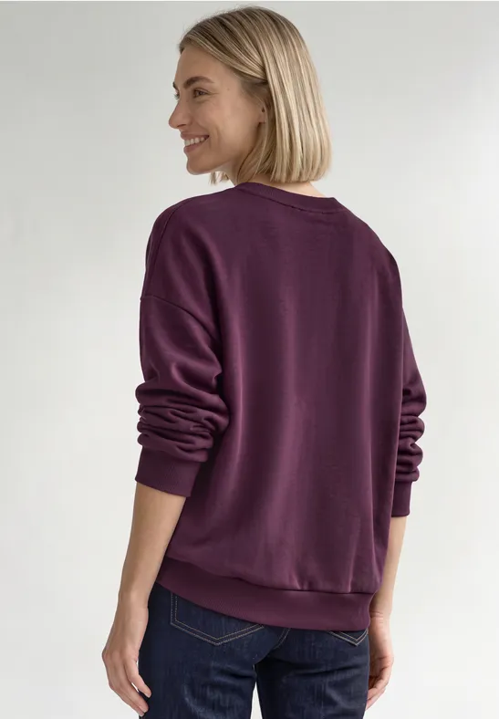 Frau von hinten mit hellblondem Bob, die ein violettes Sweatshirt trägt und lächelnd seitlich schaut.