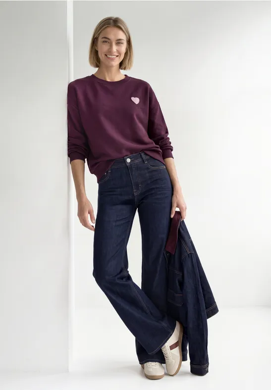 Frau in lila Pullover, weiter Jeans, weißen Sandalen, hält Jeansjacke und lächelt.