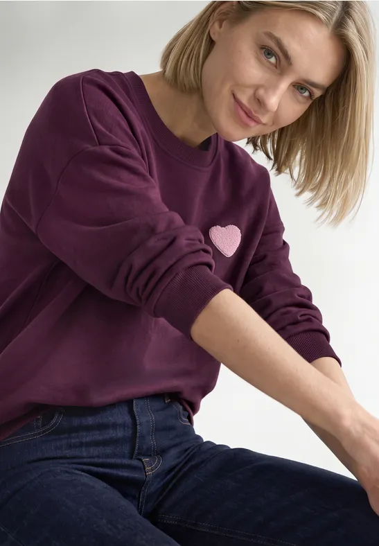 Eine lächelnde Frau in lila Sweatshirt mit Herzmotiv und blauen Jeans vor neutralem Hintergrund.