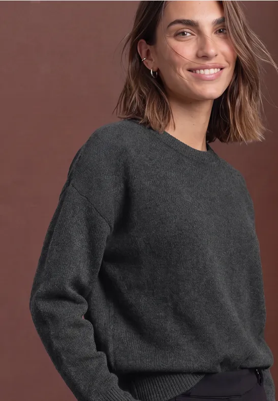 Eine lächelnde Frau in grauem Pullover vor braunem Hintergrund.