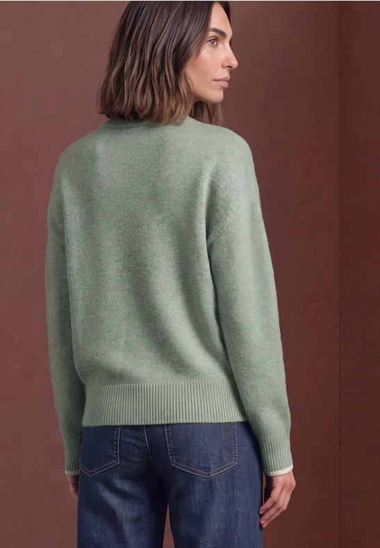 Frau von hinten in grünem Pullover und blauen Jeans vor braunem Hintergrund.