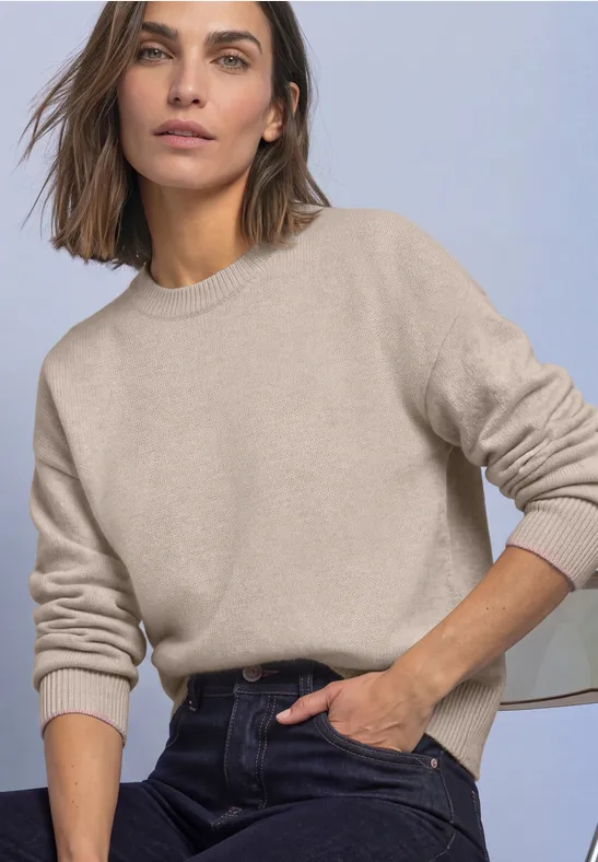 Frau in beigem Pullover und dunklen Jeans, eine Hand an der Hüfte vor hellem Hintergrund.