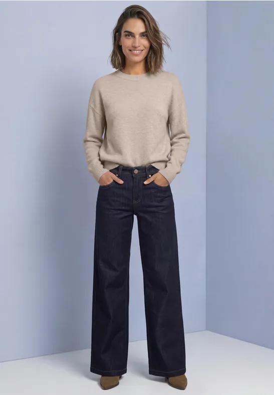 Frau mit beigem Pullover und weit geschnittenen dunklen Jeans, Hände in den Taschen, vor hellblauem Hintergrund.
