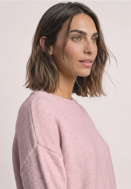 Porträt einer Frau mit braunen Haaren in rosa Pullover vor hellem Hintergrund, Blick seitlich rechts.