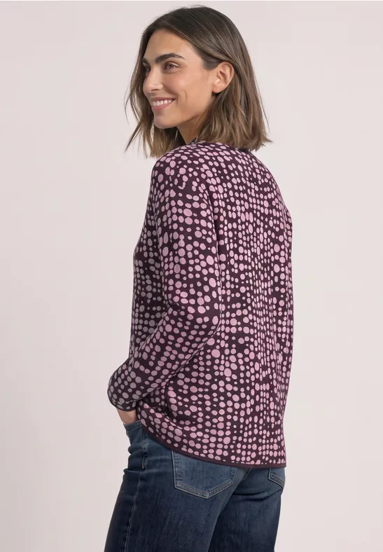 Frau im lässigen Look: gemusterte rosa-schwarze Bluse und Jeans, lächelt über die Schulter.