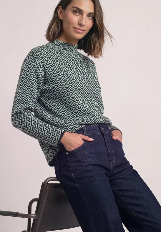 Frau in gemustertem Pullover und Jeans lehnt an einem Stuhl.