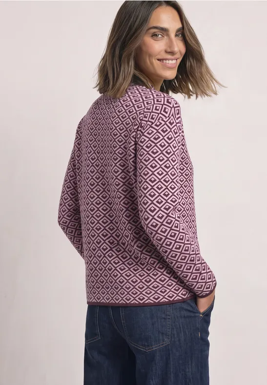 Eine Frau lächelt seitlich, trägt eine flieder-weiß gemusterte Strickjacke und blaue Jeans.