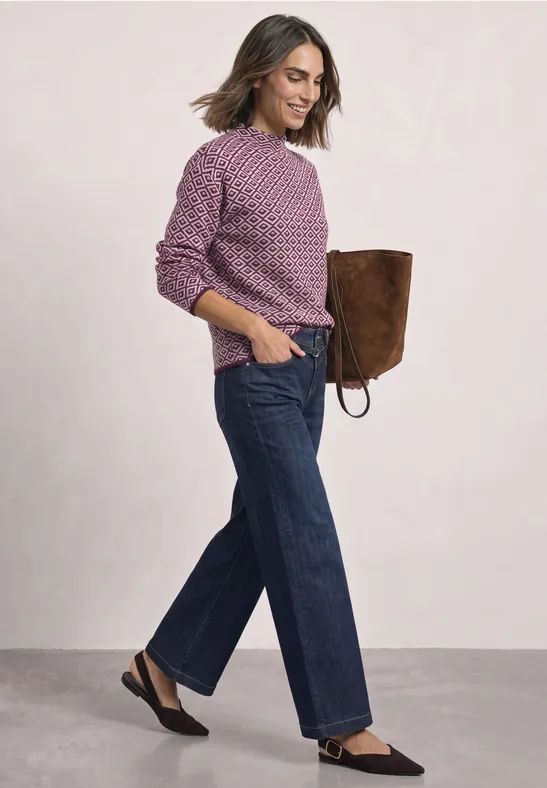 Frau in lila Strickpullover, weiten Jeans, schwarzen Slippern und brauner Tasche.