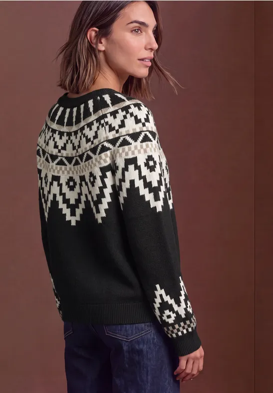 Eine Frau trägt einen schwarz-weißen Norwegerpullover und Jeans, schaut seitlich, vor braunem Hintergrund.