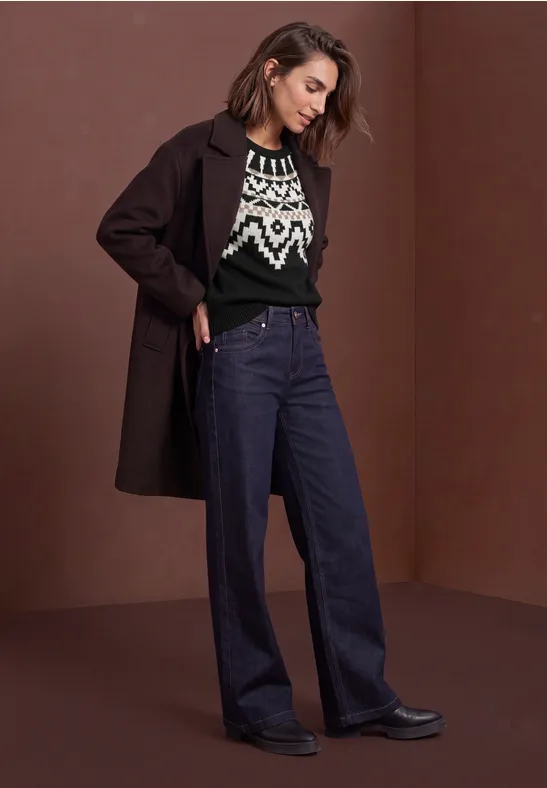 Frau in langem Mantel, gemustertem Pullover und weiten Jeans vor schlichtem braunem Hintergrund