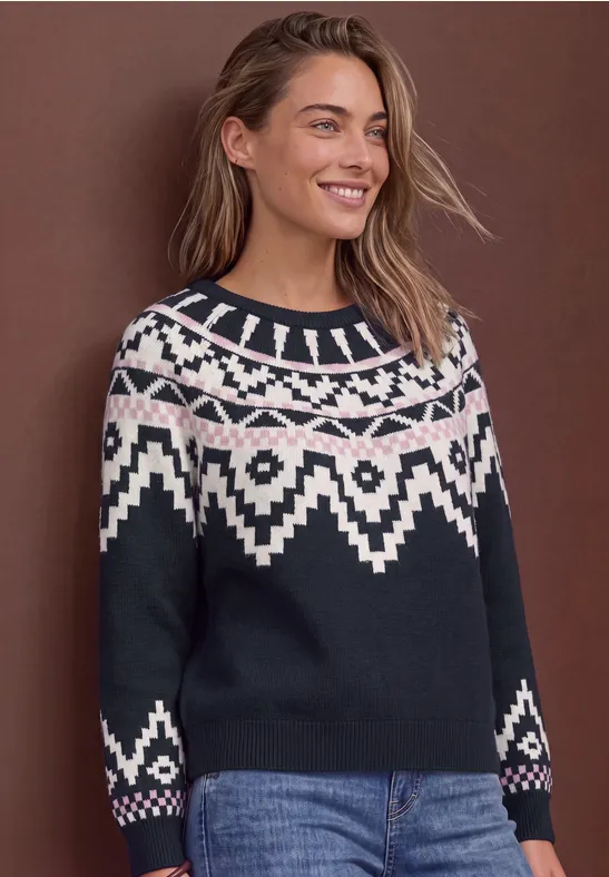 Frau in schwarzem Pullover mit weißem Norweger-Muster, Jeans, lächelnd vor braunem Hintergrund.