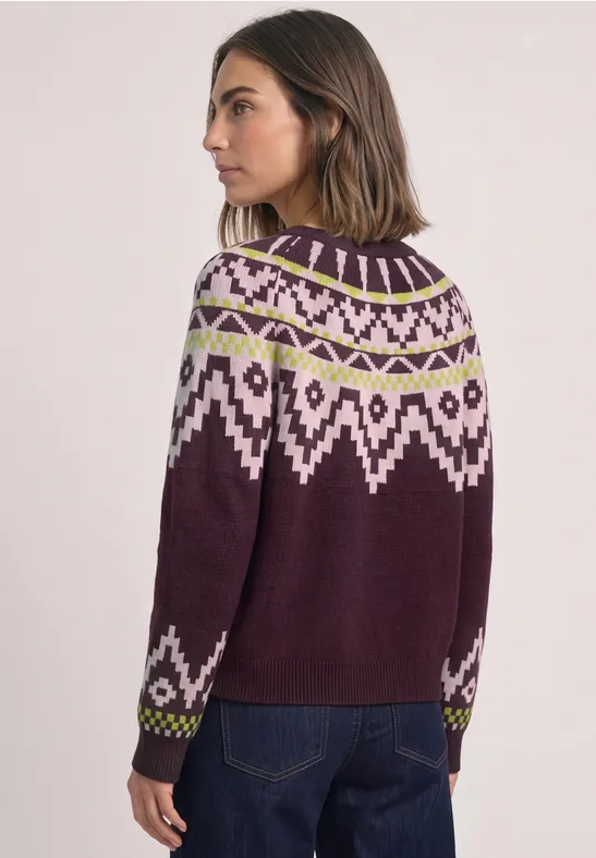 Frau trägt violetten Norwegerpullover mit weiß-grünem Muster, steht seitlich in Jeans vor hellem Hintergrund.