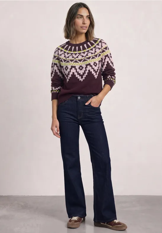 Frau in gemustertem Strickpullover und weiten Jeans steht vor hellem Hintergrund.