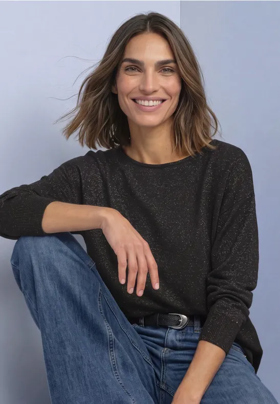 Eine lächelnde Frau mit braunen Haaren in dunkel meliertem Pullover und blauen Jeans sitzt entspannt.