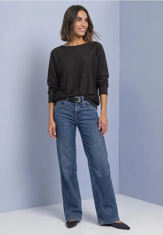 Frau in schwarzem Pulli und hoch taillierter, ausgestellter Jeans steht vor hellem Hintergrund, lächelnd.