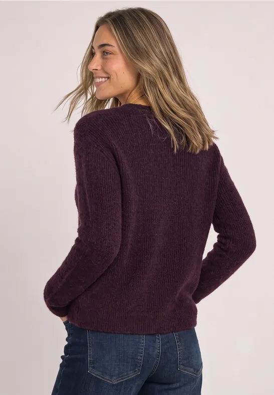 Frau mit dunkellila Strickpullover und Jeans, schaut lächelnd über die Schulter vor hellem Hintergrund.