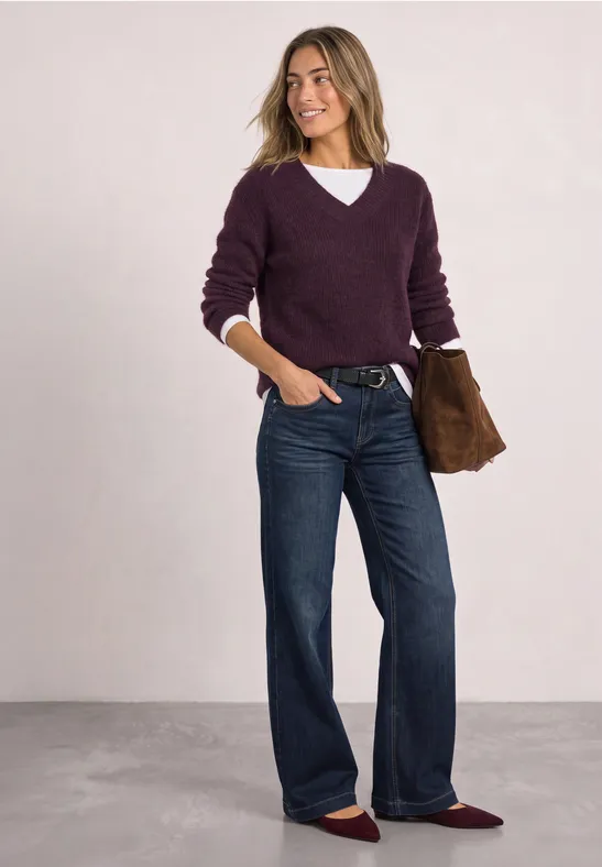 Frau in violettem Pullover, weitem Jeans, weinroten Ballerinas und Clutch.