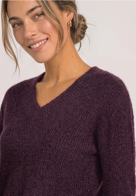 Frau mit blondem Dutt trägt violetten Strickpullover mit V-Ausschnitt und lächelt.