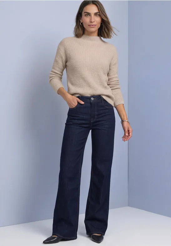 Frau in beigem Pullover und weitem, hoch tailliertem dunkelblauen Jeans vor hellem Hintergrund.