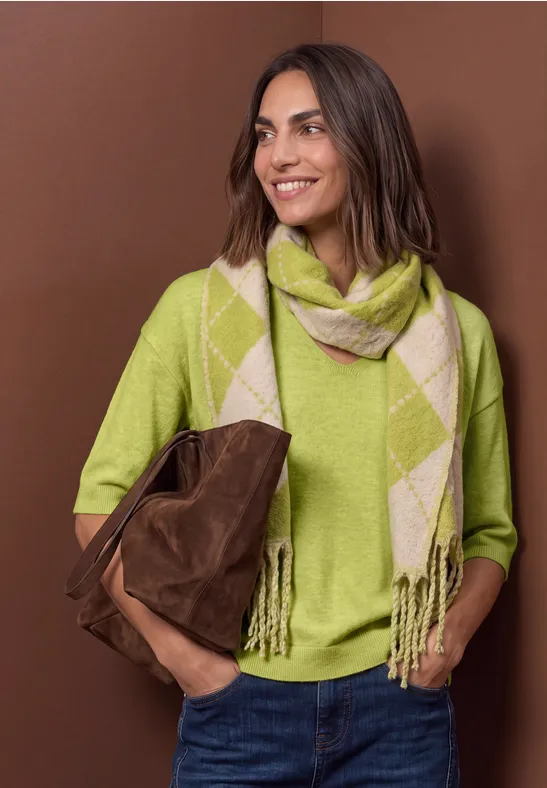 Femme souriante en pull vert et écharpe à carreaux tenant un sac marron.