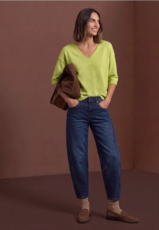 Femme en jean bleu décontracté, pull vert à col V et mocassins beiges, tenant un blazer marron.