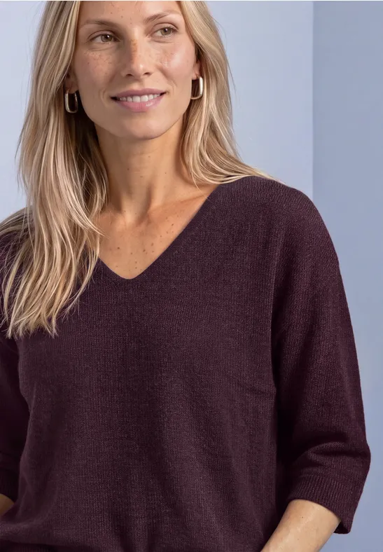 Kobieta o blond włosach ma na sobie ciemnofioletowy sweter w serek z 3/4 rękawami i kołowymi kolczykami.