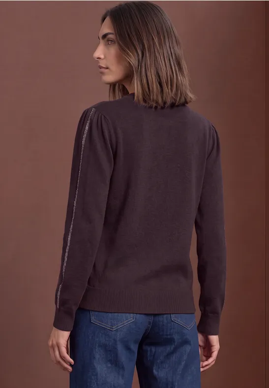 Eine Frau von hinten in Jeans und braunem Pullover vor neutralem Hintergrund.
