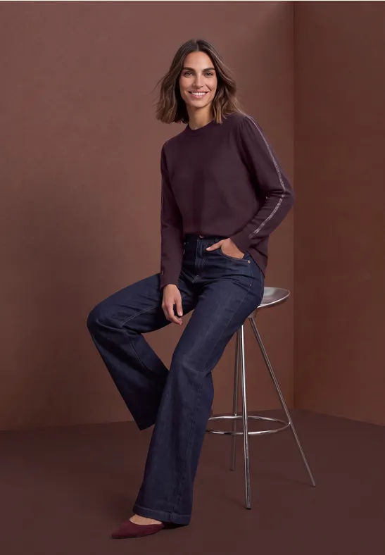 Frau in lila Pullover und weiten Jeans sitzt lächelnd auf einem Hocker vor braunem Hintergrund.