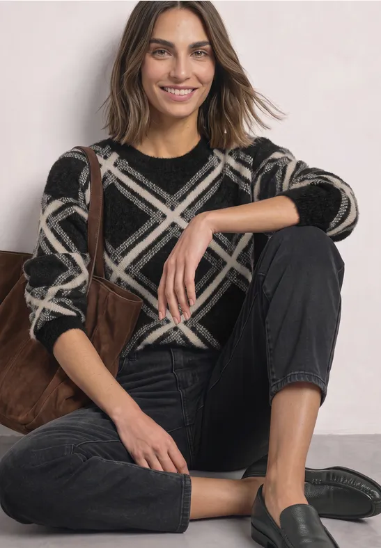 Frau sitzt lächelnd auf dem Boden, trägt schwarzen Pullover mit weißem Karomuster, schwarze Jeans und Loafers.