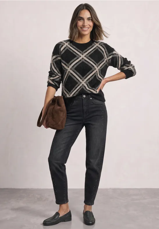 Frau in schwarzer Jeans und gemustertem Pullover mit brauner Tasche und Loafers vor hellem Hintergrund.