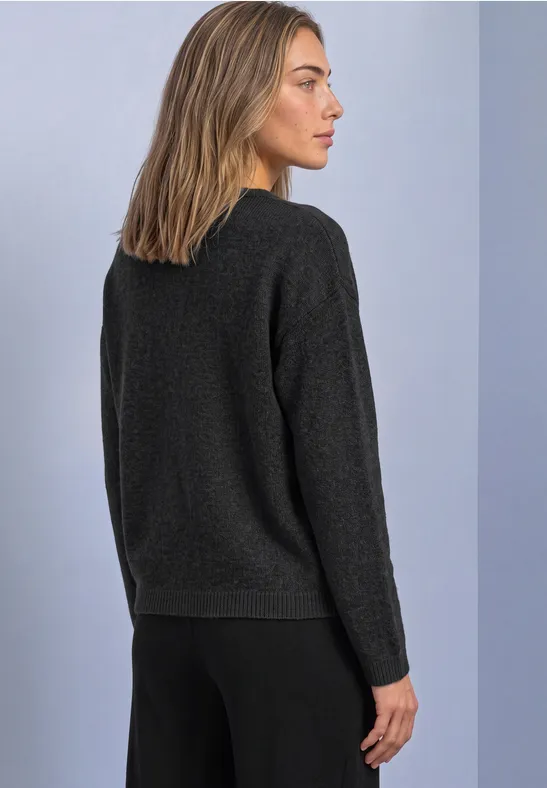 Seitliche Ansicht einer Frau in schwarzem Pullover und Hose, vor hellem Hintergrund.