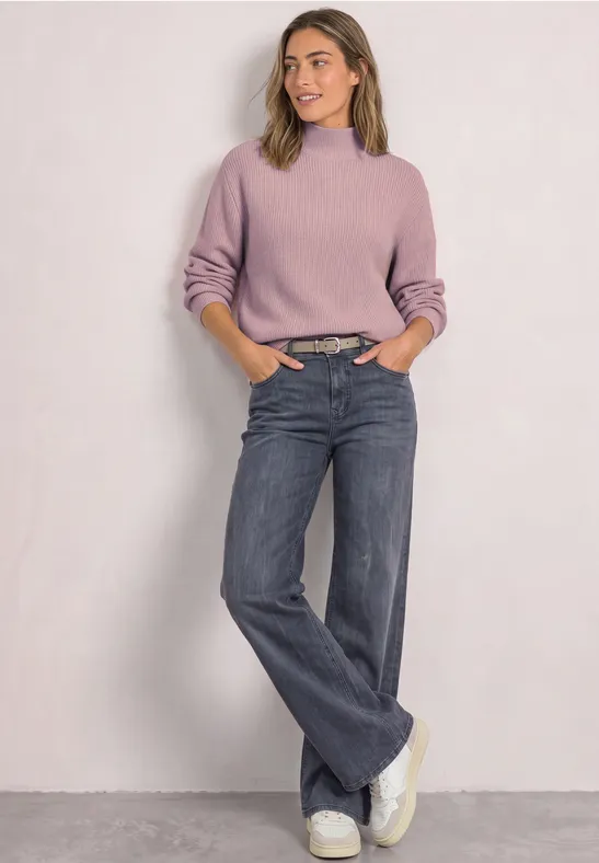 Frau in fliederfarbenem Pullover, weiten Jeans und weißen Sneakern vor schlichtem Hintergrund.