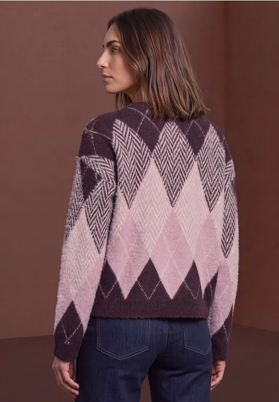 Frau steht seitlich, trägt violett-rosa Argyle-Pullover und Jeans vor braunem Hintergrund.