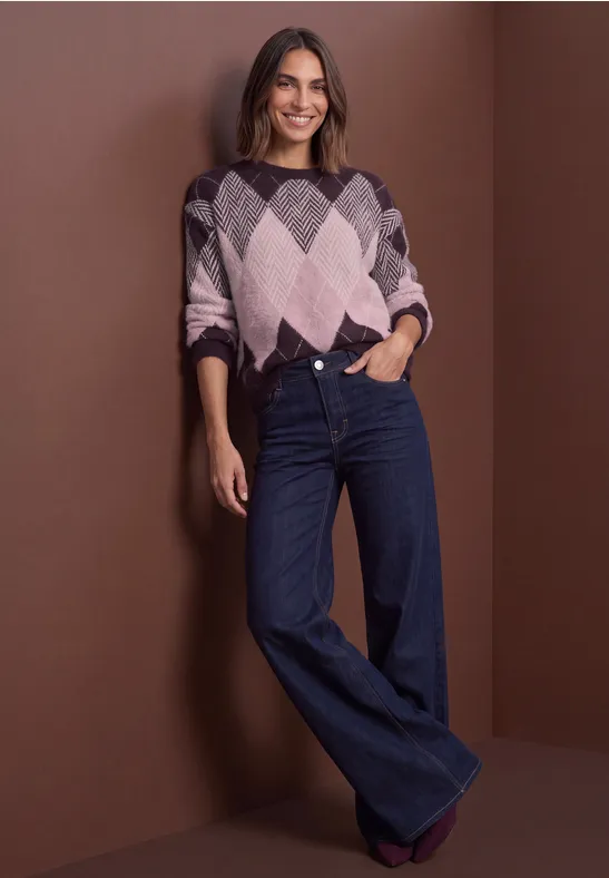 Frau mit bunt gestricktem Pullover und weiten Jeans lehnt lächelnd an einer braunen Wand.