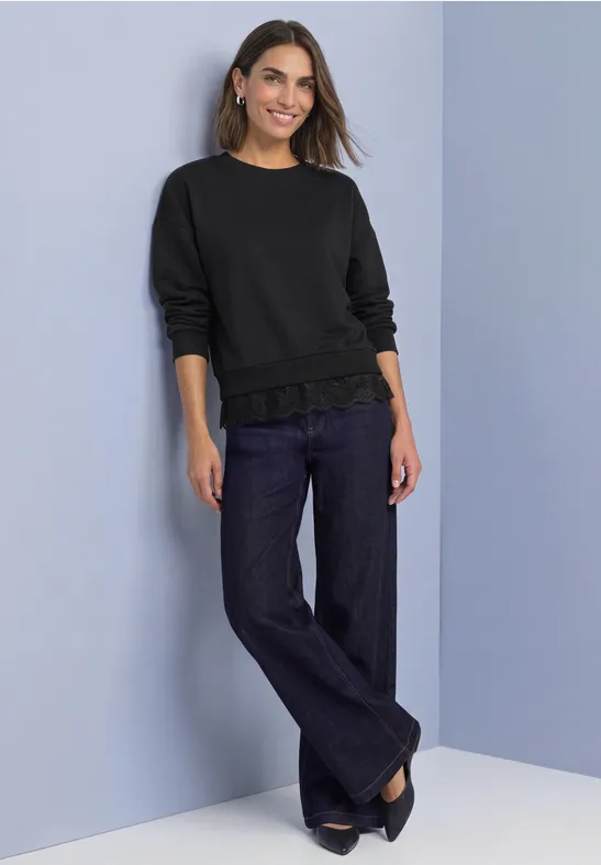 Frau in schwarzem Pullover und weitem Jeans, lächelt vor hellblauem Hintergrund.